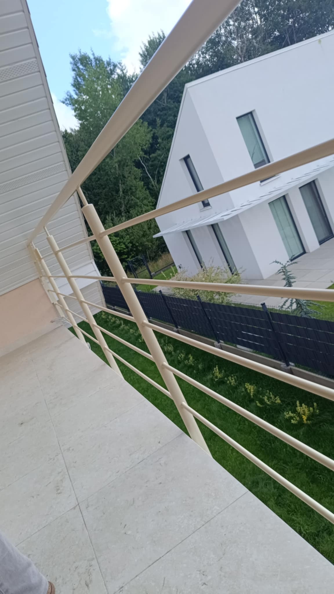 Biała, metalowa balustrada balkonowa z kremowymi słupkami na tle nowoczesnego domu z dużymi oknami i zielonego trawnika. Ujęcie z dołu, lekko pod kątem.