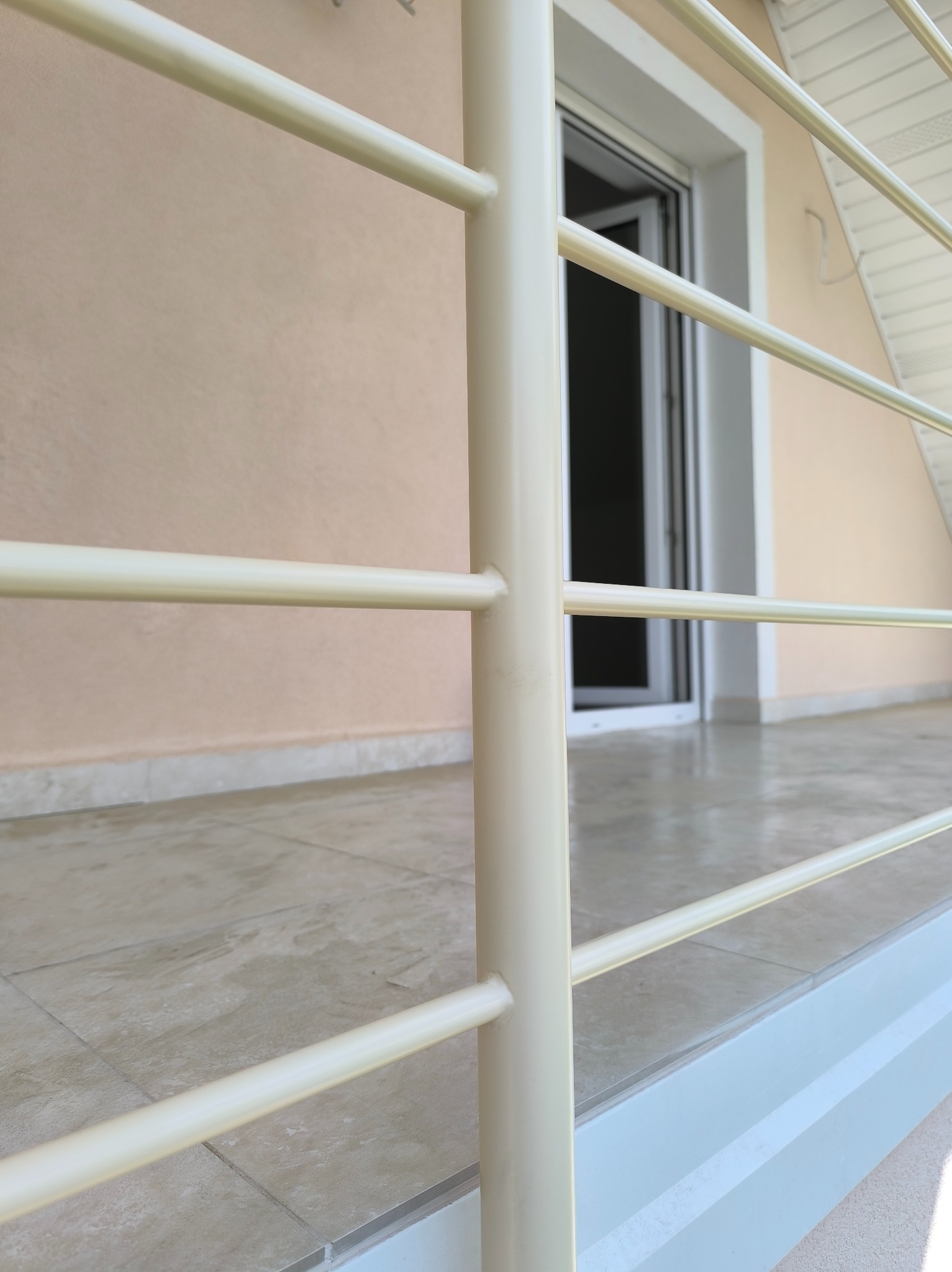 Jasna, metalowa balustrada balkonowa z poziomymi prętami, widok z dołu, z oknem balkonowym w tle i beżową ścianą budynku. Detal łączenia prętów.