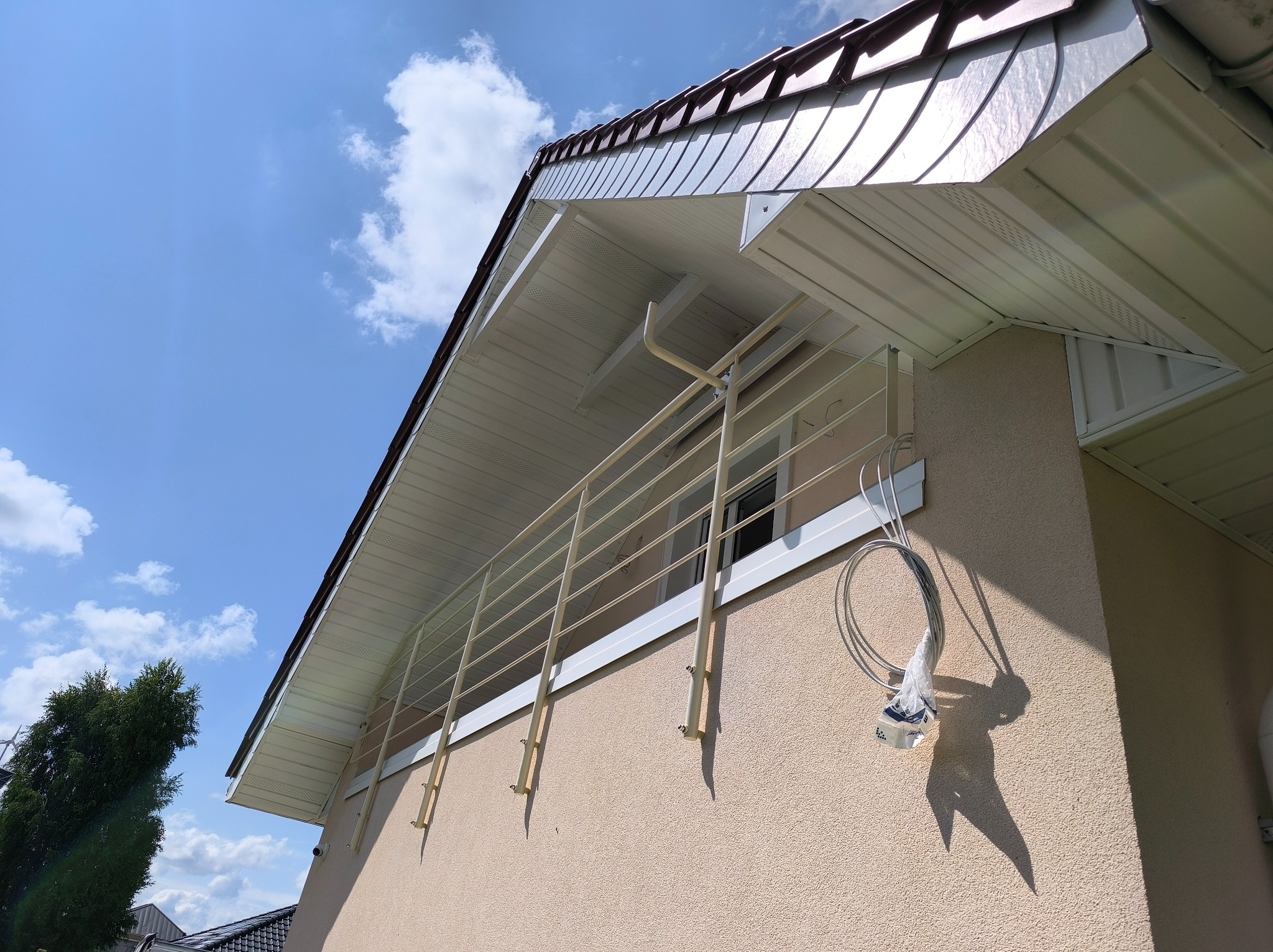 Balkon z kremową, metalową konstrukcją w Przećminie, widziany z dołu pod kątem. Widoczne błękitne niebo i fragment dachu budynku. Na ścianie zwisają kable.