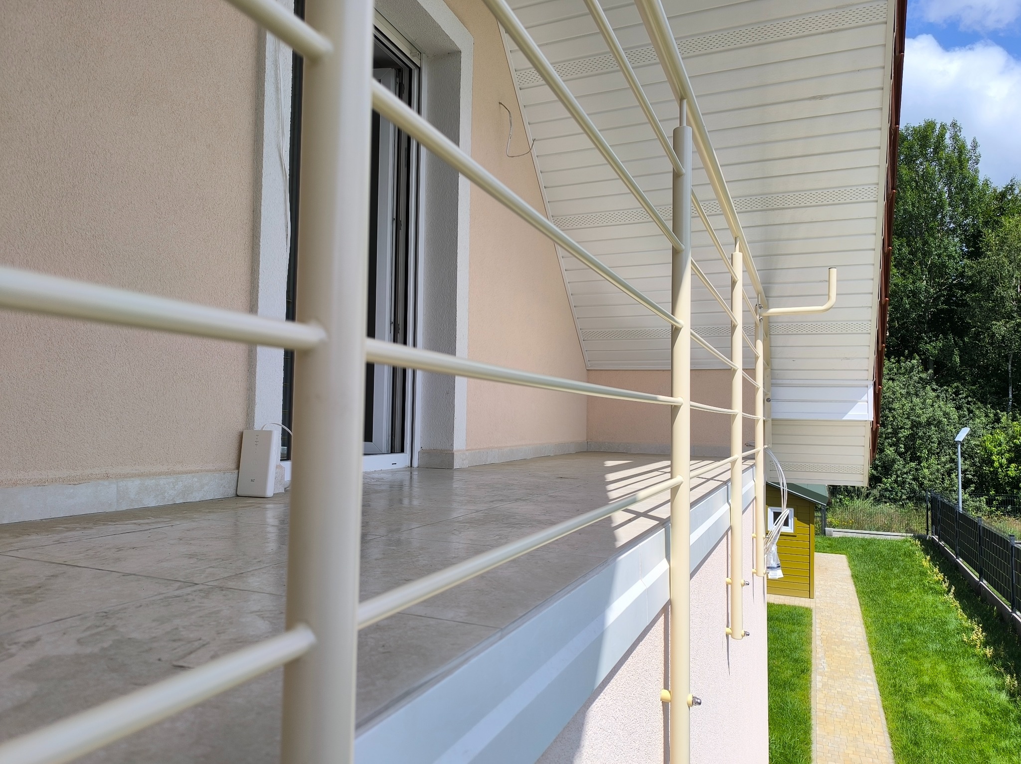 Jasna, metalowa balustrada balkonowa z poziomymi poprzeczkami, widok z boku, z fragmentem budynku i zieleni w tle, w słoneczny dzień.