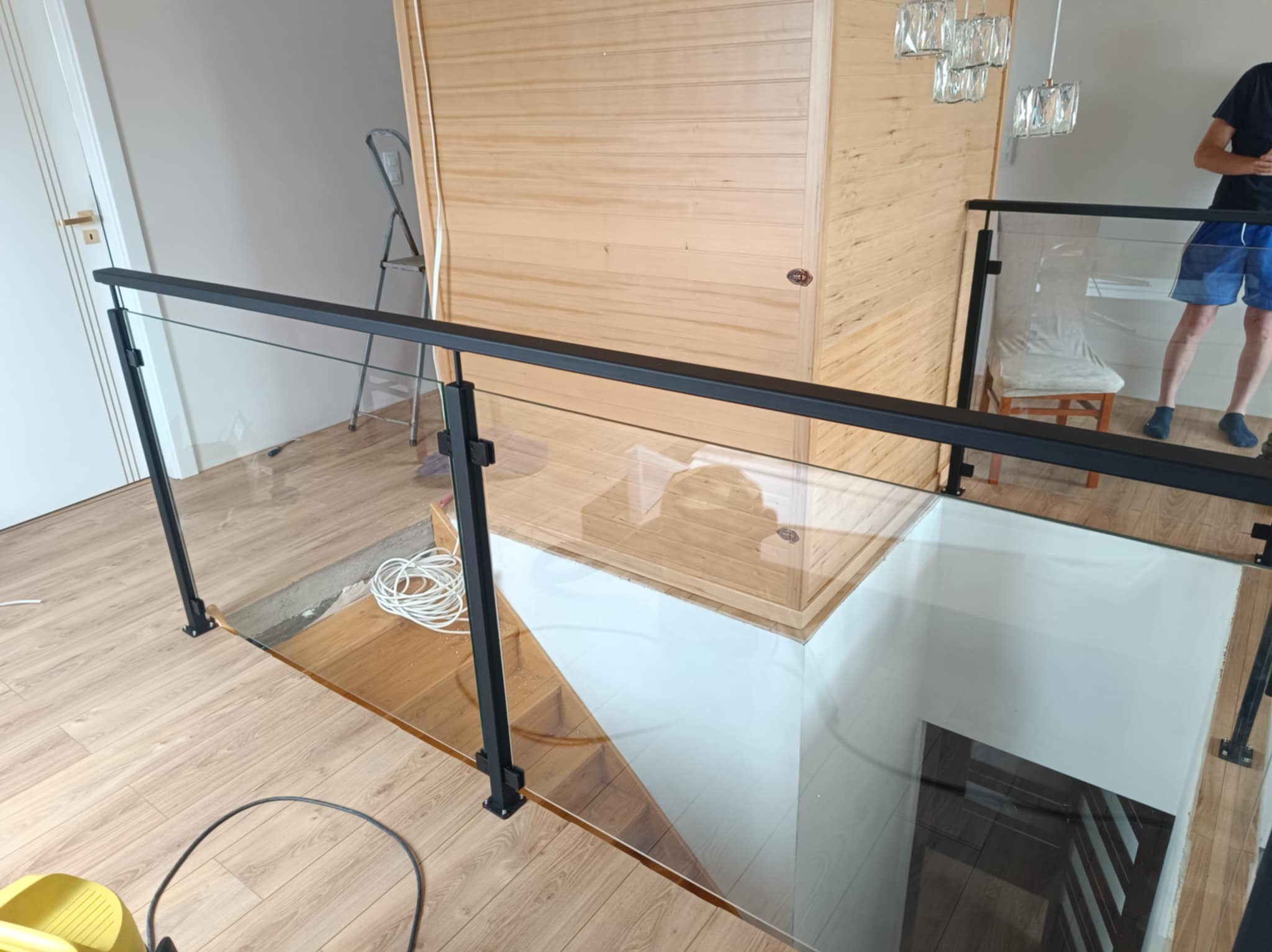 Szklana balustrada z czarnymi elementami mocującymi zabezpiecza schody. Widoczne drewniane wykończenie podłogi i schodów. W tle drzwi i drabina.