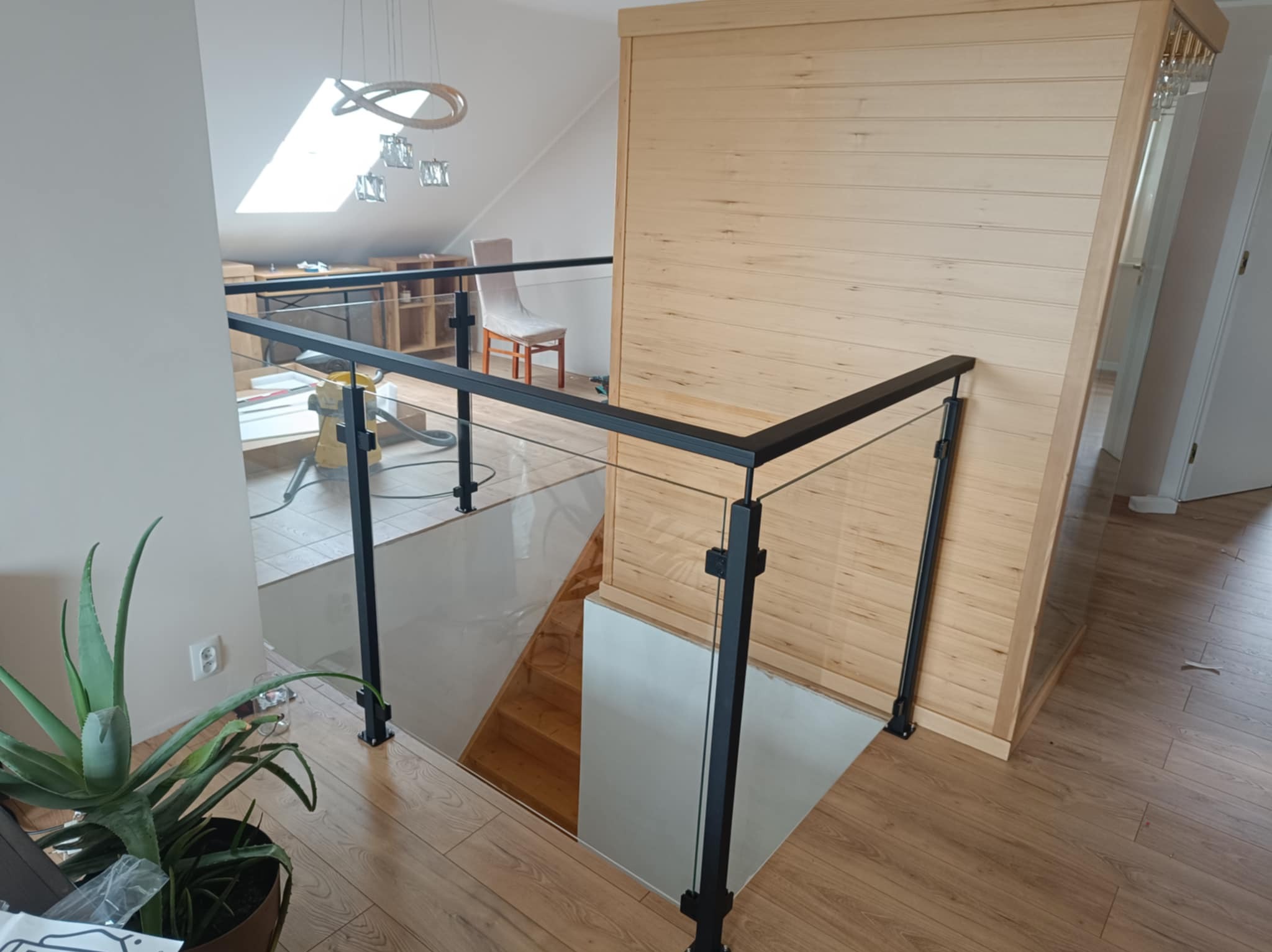 Szklana balustrada z czarnymi, metalowymi słupkami w nowoczesnym wnętrzu. Widoczny fragment schodów, drewniana boazeria i roślina doniczkowa.