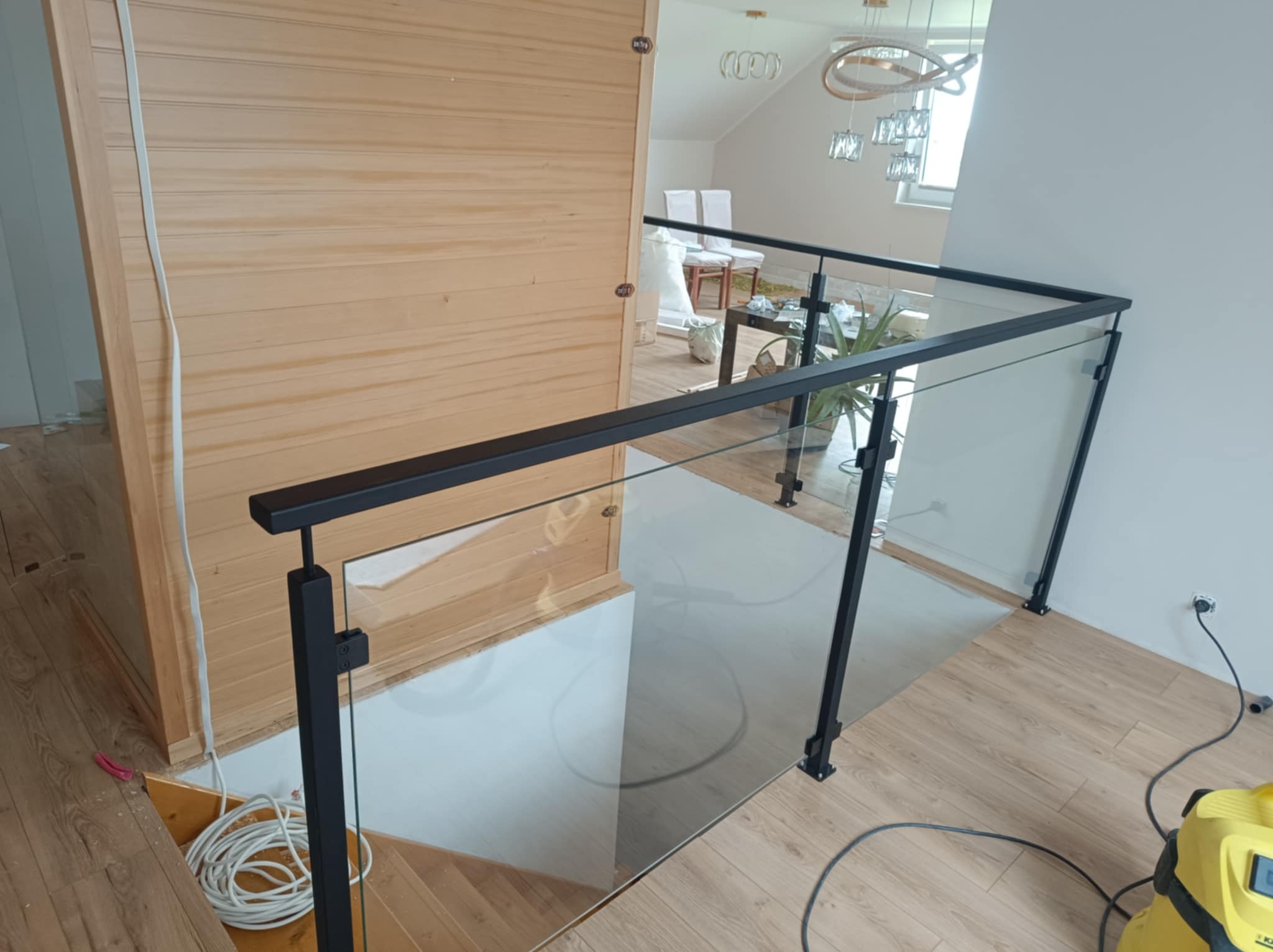 Nowoczesna balustrada szklana z czarnymi elementami metalowymi, zamontowana na schodach wewnątrz domu. Widoczne drewniane wykończenia i jasne wnętrze.