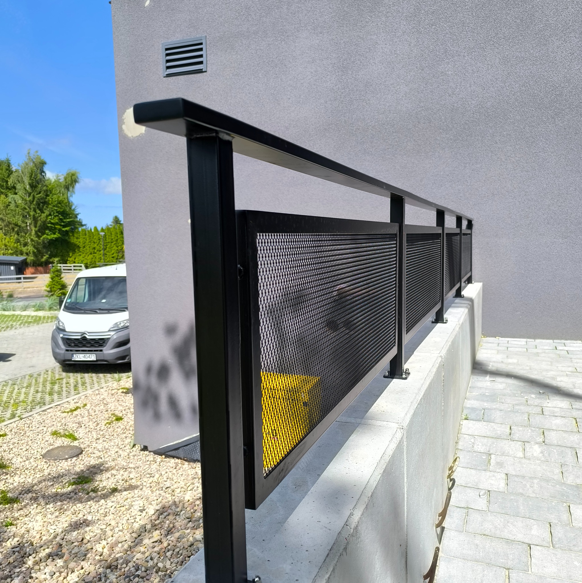 Czarna, metalowa poręcz z perforowanym wypełnieniem, zamontowana na betonowym murku przy schodach wejściowych do budynku. W tle widoczny samochód dostawczy.