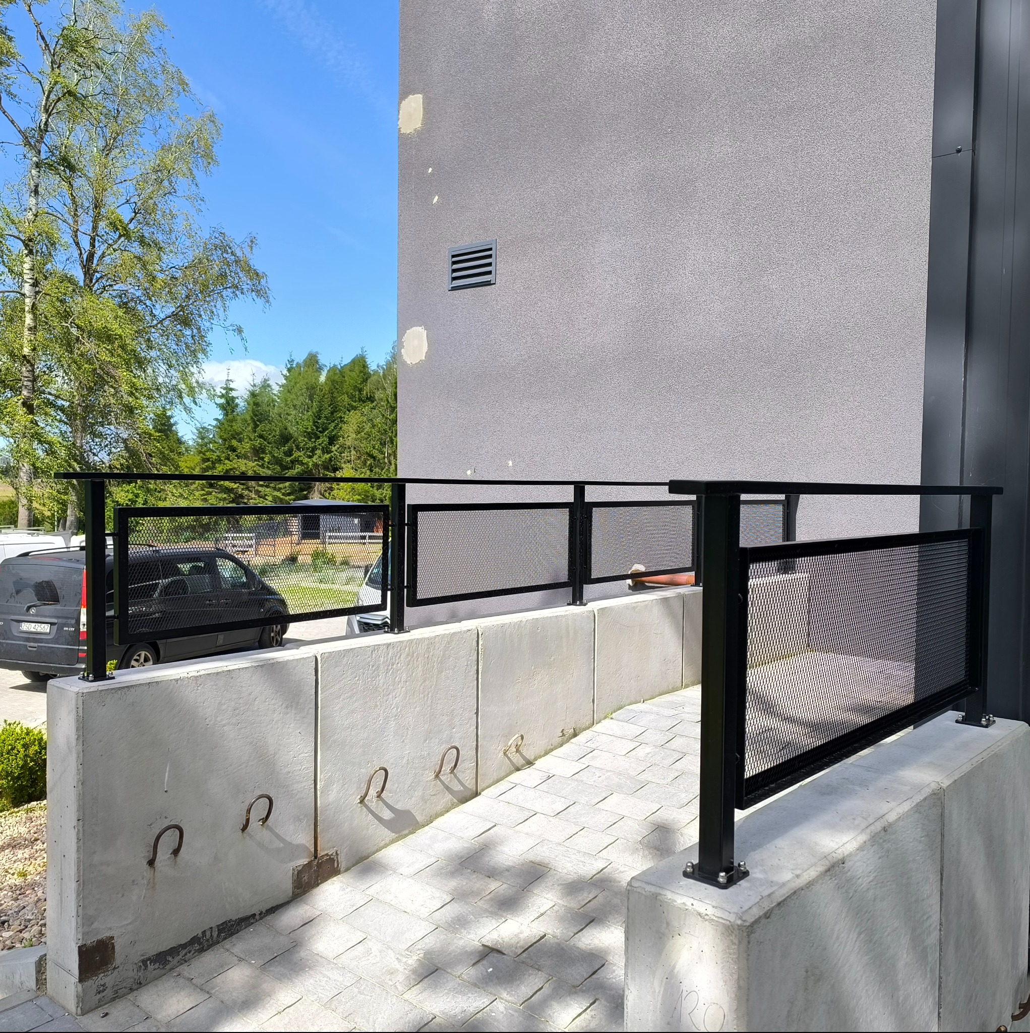 Czarna, metalowa balustrada z siatkowym wypełnieniem, zamontowana na betonowym murku. W tle budynek z szarą elewacją i fragment zieleni.