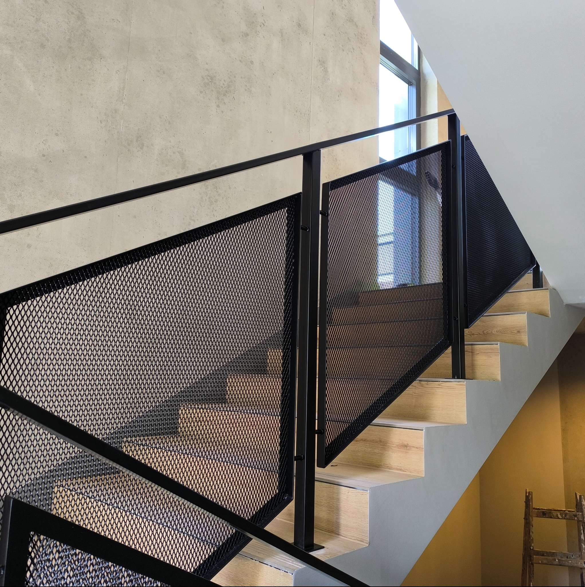 Nowoczesne schody z drewnianymi stopniami i czarną, metalową balustradą z siatki, widziane z dołu, z fragmentem okna w tle. Minimalistyczny design.