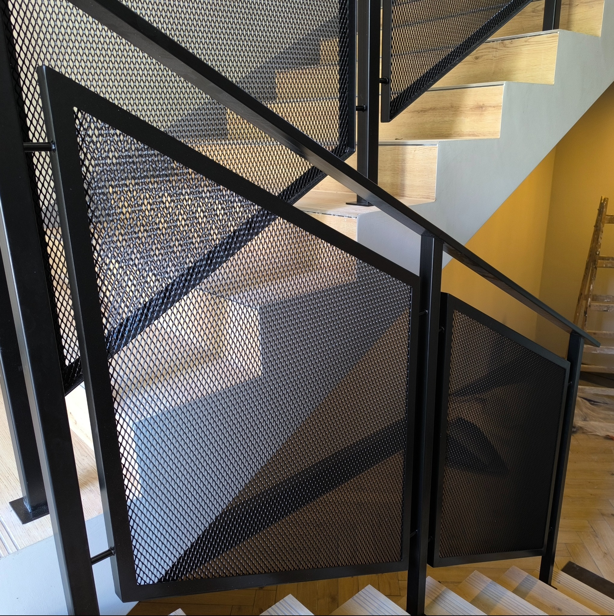 Nowoczesne schody z czarną, metalową balustradą z siatki o romboidalnych oczkach. Jasne, drewniane stopnie. Ujęcie z dołu, podkreślające geometrię konstrukcji.