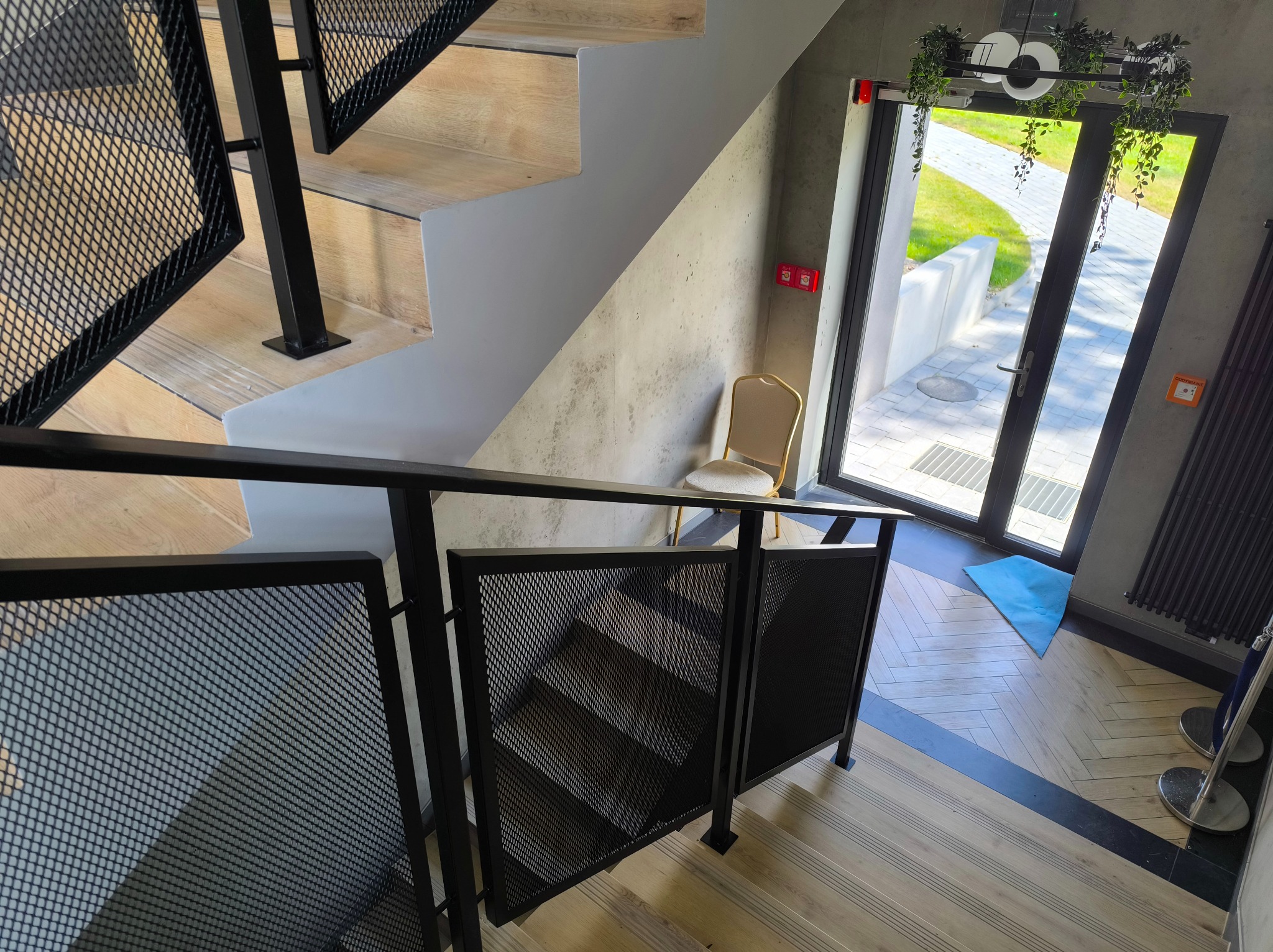 Nowoczesne schody z drewnianymi stopniami i czarną, metalową balustradą z siatki, widziane z góry. W tle jasne wnętrze z krzesłem i wyjściem na zewnątrz.
