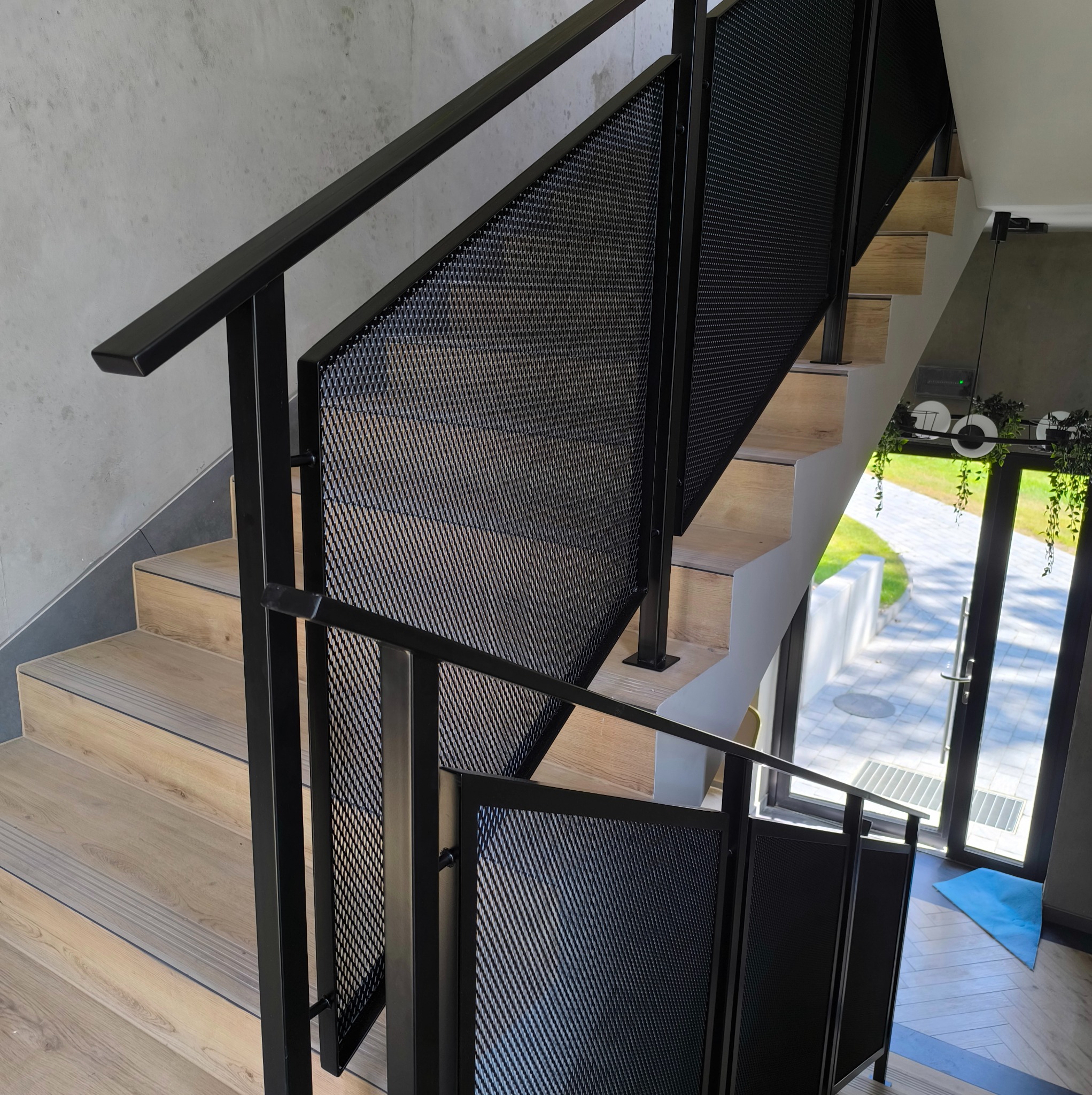 Schody z drewnianymi stopniami i czarną, metalową balustradą z siatkowym wypełnieniem. Widok na drzwi wejściowe i zewnętrzny chodnik. Nowoczesny design.