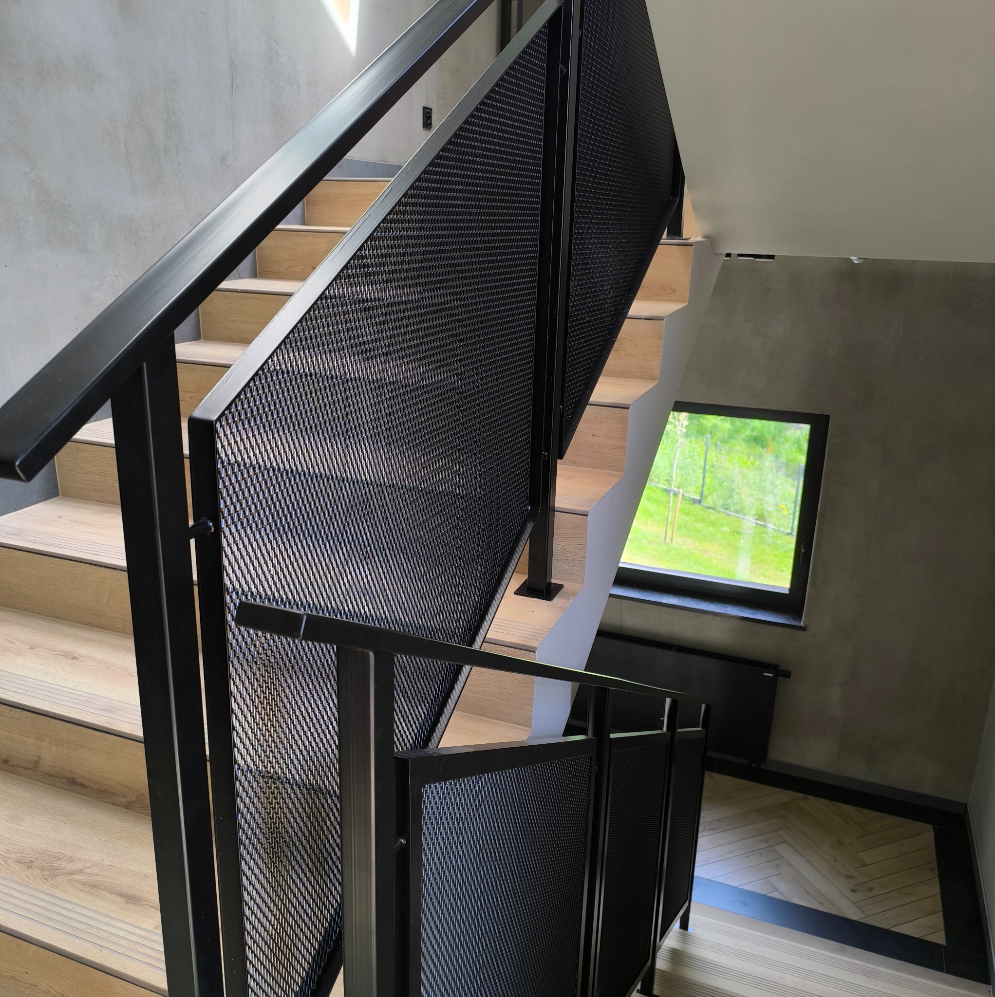 Nowoczesne schody z drewnianymi stopniami i czarną metalową balustradą z perforowanej blachy, oświetlone naturalnym światłem z okna.