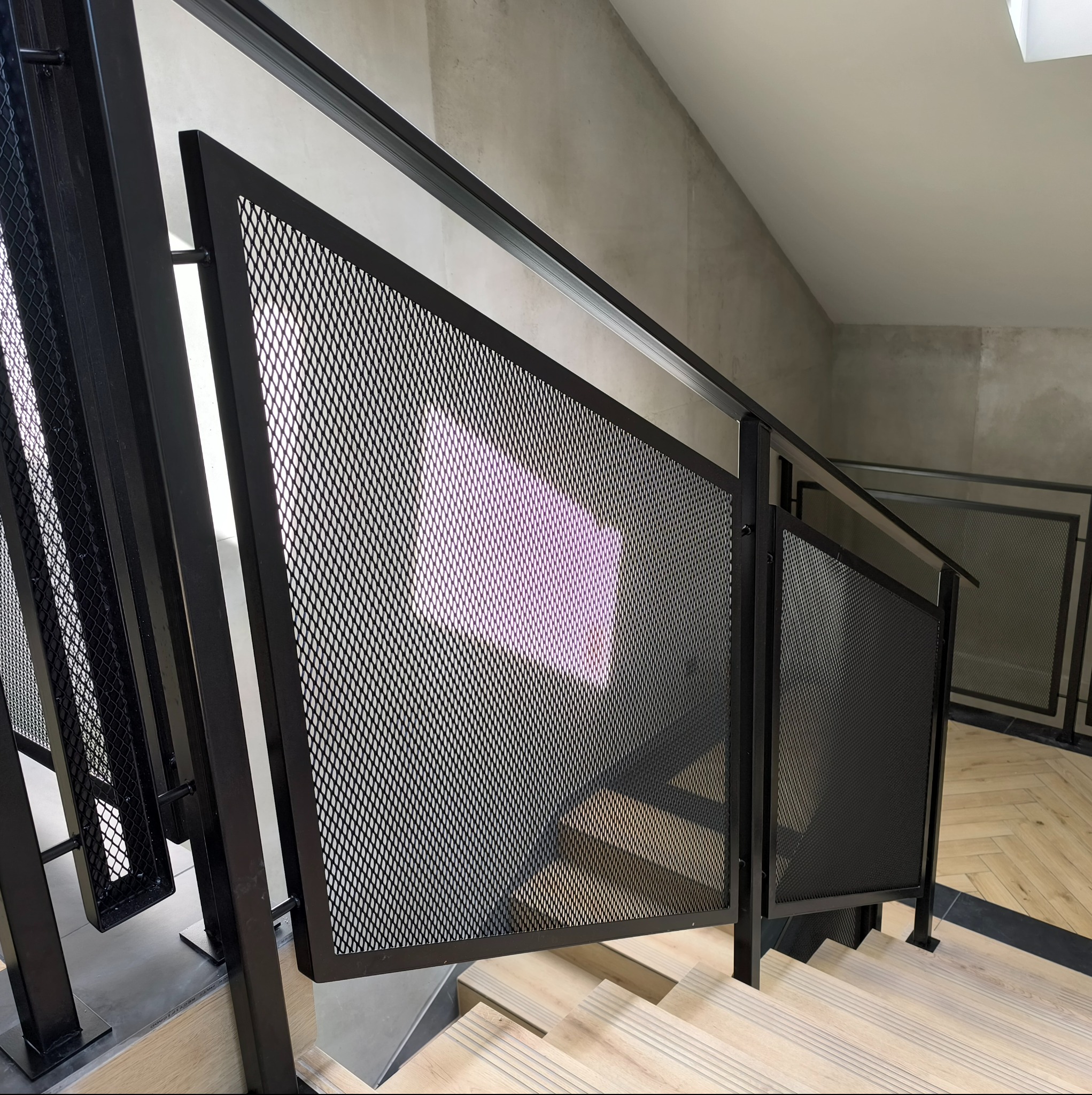 Metalowa balustrada schodowa z siatką w czarnym kolorze, widok z góry na jasne drewniane schody. Minimalistyczny design z surowym betonem w tle.