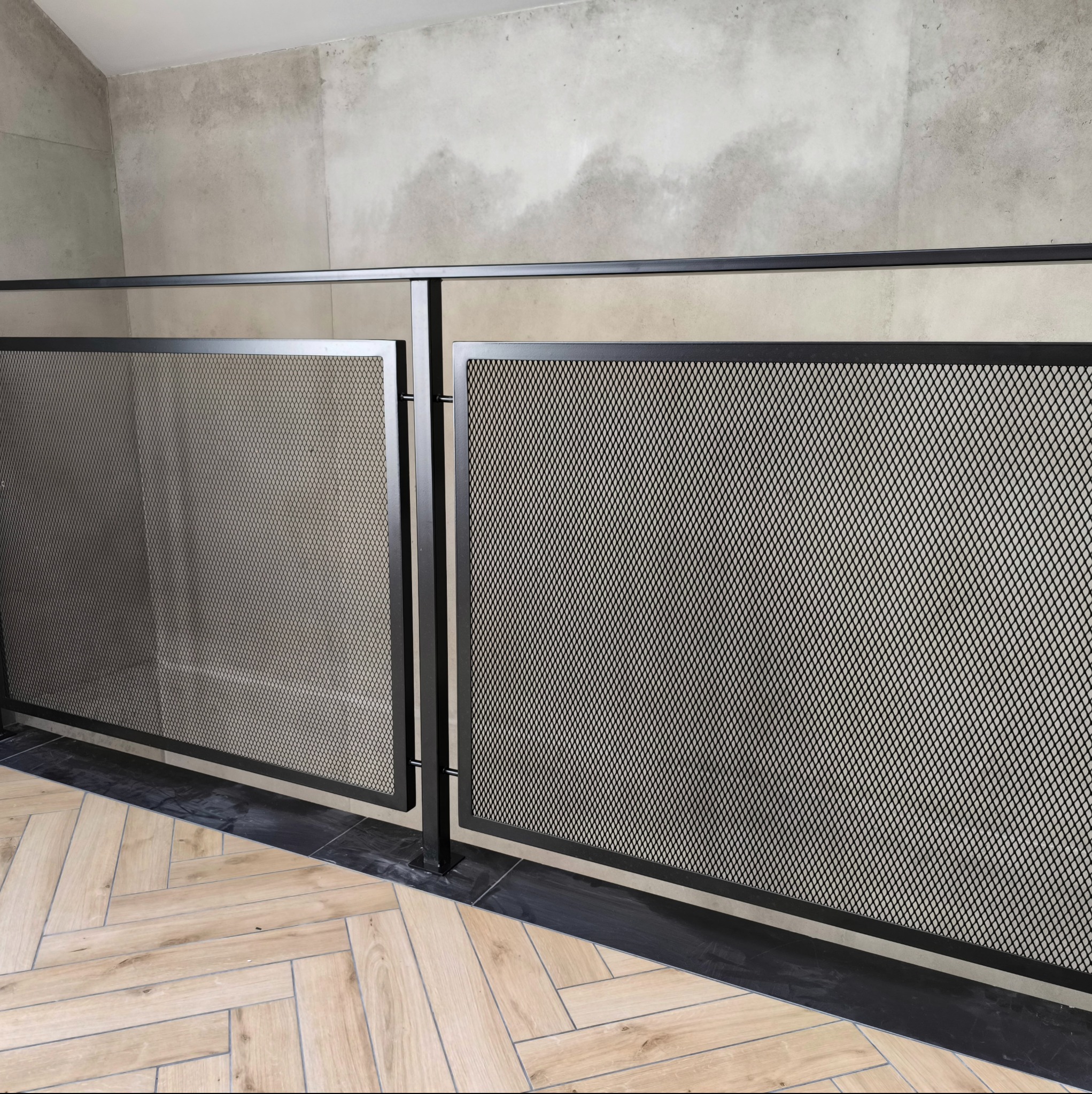 Nowoczesna, czarna balustrada z siatki o romboidalnych oczkach, zamontowana wewnątrz budynku. Widoczna podłoga z paneli ułożonych w jodełkę i szare ściany.