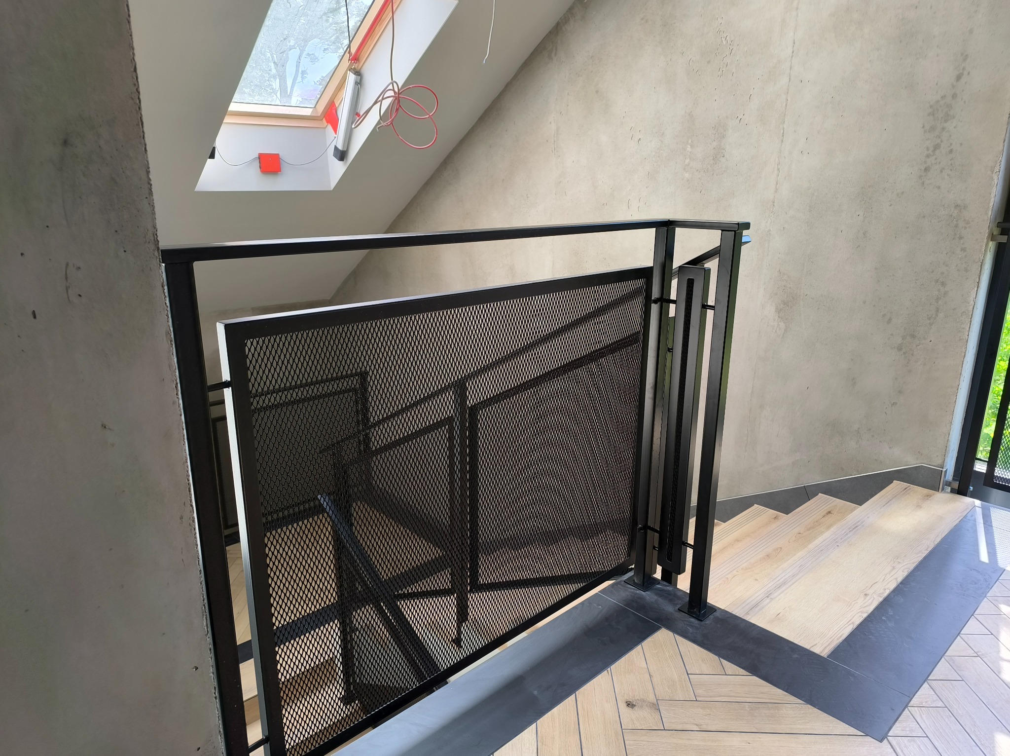 Metalowa balustrada schodowa z siatkowym wypełnieniem, czarne wykończenie, widok z góry na drewniane stopnie z szarymi akcentami, surowe betonowe ściany, doświetlenie z okna dachowego.