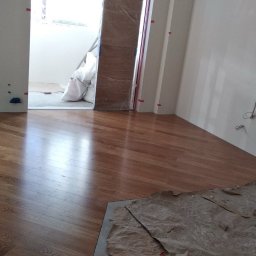 VVB group PL - Wnętrze w trakcie prac wykończeniowych: ułożona drewniana podłoga z widocznymi pozostałościami po pracach malarskich i wiaderkami z farbą, ściany przygotowane do malowania, odkryte instalacje...