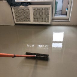 VVB group PL - Aplikacja jasnoszarej posadzki żywicznej za pomocą wałka kolczastego w pomieszczeniu z grzejnikiem pod oknem, widoczne odbicie światła w mokrej powierzchni.