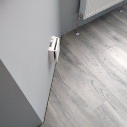 VVB group PL - Szare panele podłogowe ułożone wzdłuż ściany z wystającym elementem instalacji elektrycznej oraz grzejnikiem w tle.