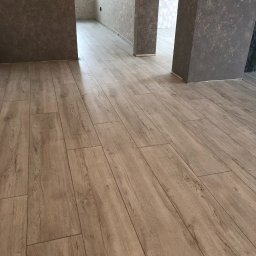 VVB group PL - Jasne panele podłogowe imitujące drewno, ułożone wzdłuż korytarza z szarobrązowymi ścianami o nierównej fakturze, widok w kierunku dwóch otwartych przejść do innych pomieszczeń.