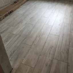 Ułożone panele podłogowe w jasnym odcieniu drewna, widoczne kartony z panelami przy ścianie, grzejnik na ścianie w tle.