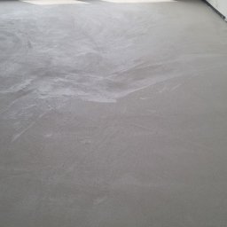 VVB group PL - Szara, gładka posadzka z widocznymi smugami i refleksami światła, w tle okno z workami gruzu na parapecie.