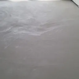 Szara, gładka posadzka z widocznymi smugami i refleksami światła, w tle okno z workami gruzu na parapecie.