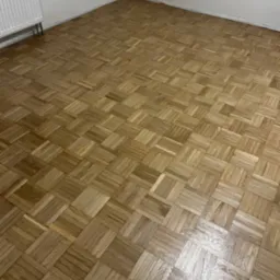 Odnowiona podłoga parkietowa w jodełkę, po cyklinowaniu i lakierowaniu. Widoczne naturalne usłojenie drewna i ciepła kolorystyka. W tle biała ściana z grzejnikiem.