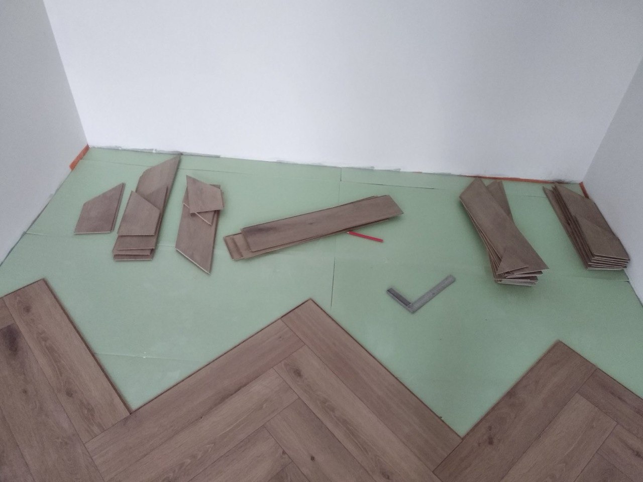 Fragment podłogi w trakcie montażu z ułożonym wzorem w jodełkę; widoczne docięte panele, zielona podkładka i kątownik.