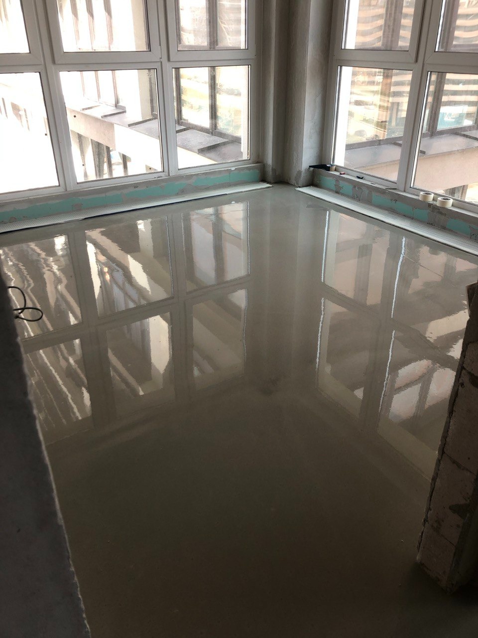 Świeżo wylana, gładka, samopoziomująca posadzka w pomieszczeniu z dużymi oknami, odbijającymi światło.