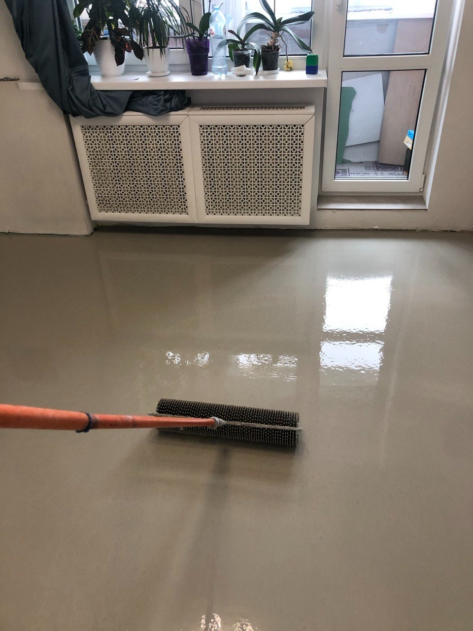 Aplikacja jasnoszarej posadzki żywicznej za pomocą wałka kolczastego w pomieszczeniu z grzejnikiem pod oknem, widoczne odbicie światła w mokrej powierzchni.