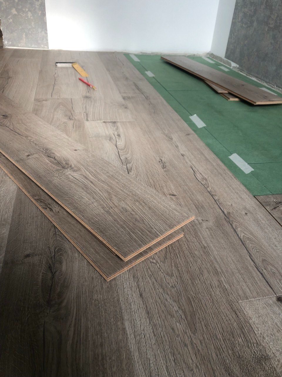 Fragment podłogi w trakcie montażu, widoczne panele o wzorze drewna, zielona mata podkładowa, ołówek i miarka, przy białej ścianie.