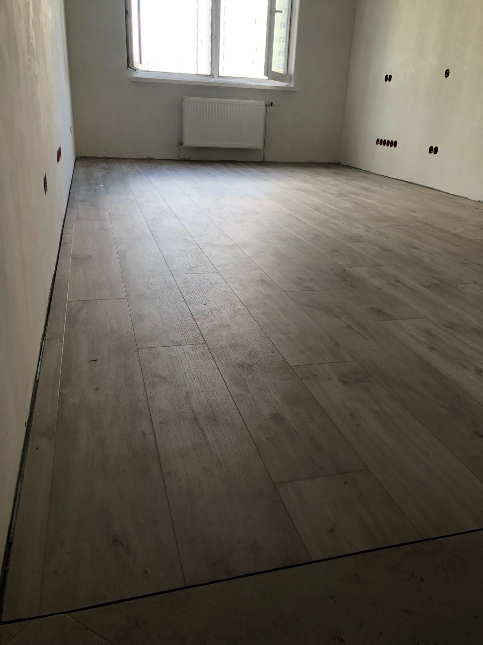 Ułożone panele podłogowe imitujące drewno w jasnym pomieszczeniu z grzejnikiem pod oknem i gniazdkami elektrycznymi na ścianie.