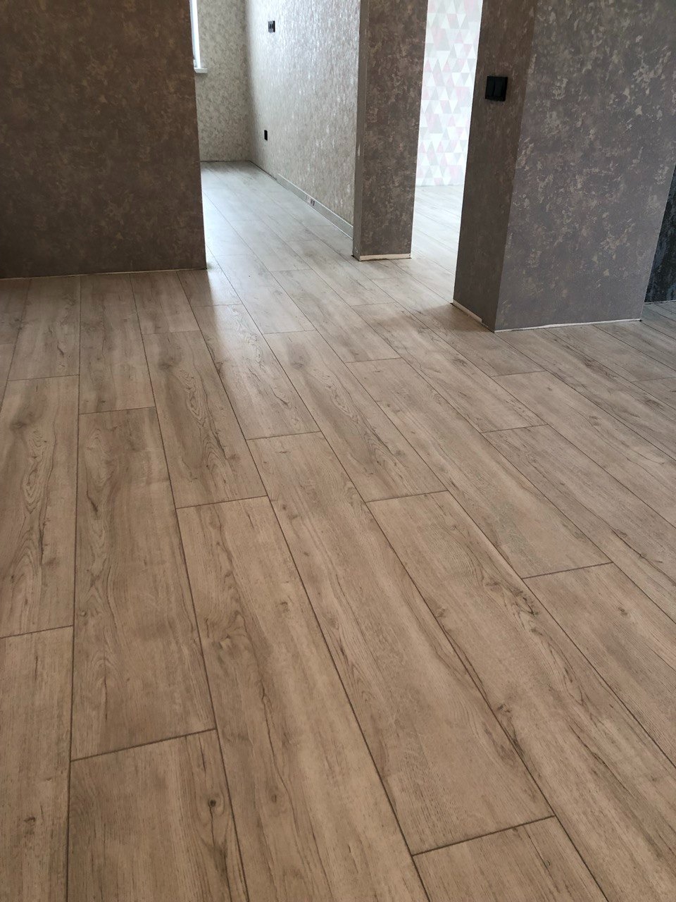 Jasne panele podłogowe imitujące drewno, ułożone wzdłuż korytarza z szarobrązowymi ścianami o nierównej fakturze, widok w kierunku dwóch otwartych przejść do innych pomieszczeń.