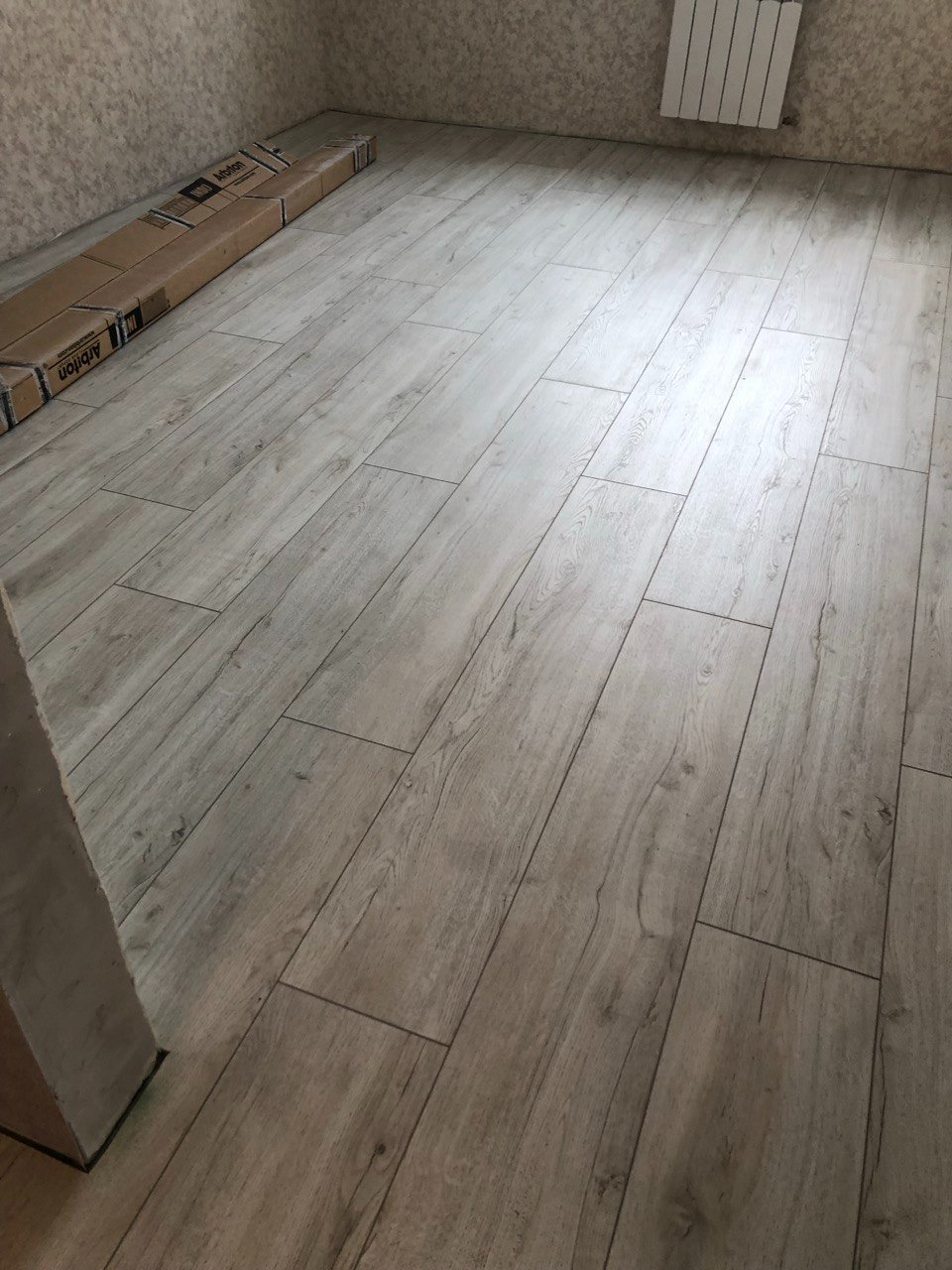 Ułożone panele podłogowe w jasnym odcieniu drewna, widoczne kartony z panelami przy ścianie, grzejnik na ścianie w tle.