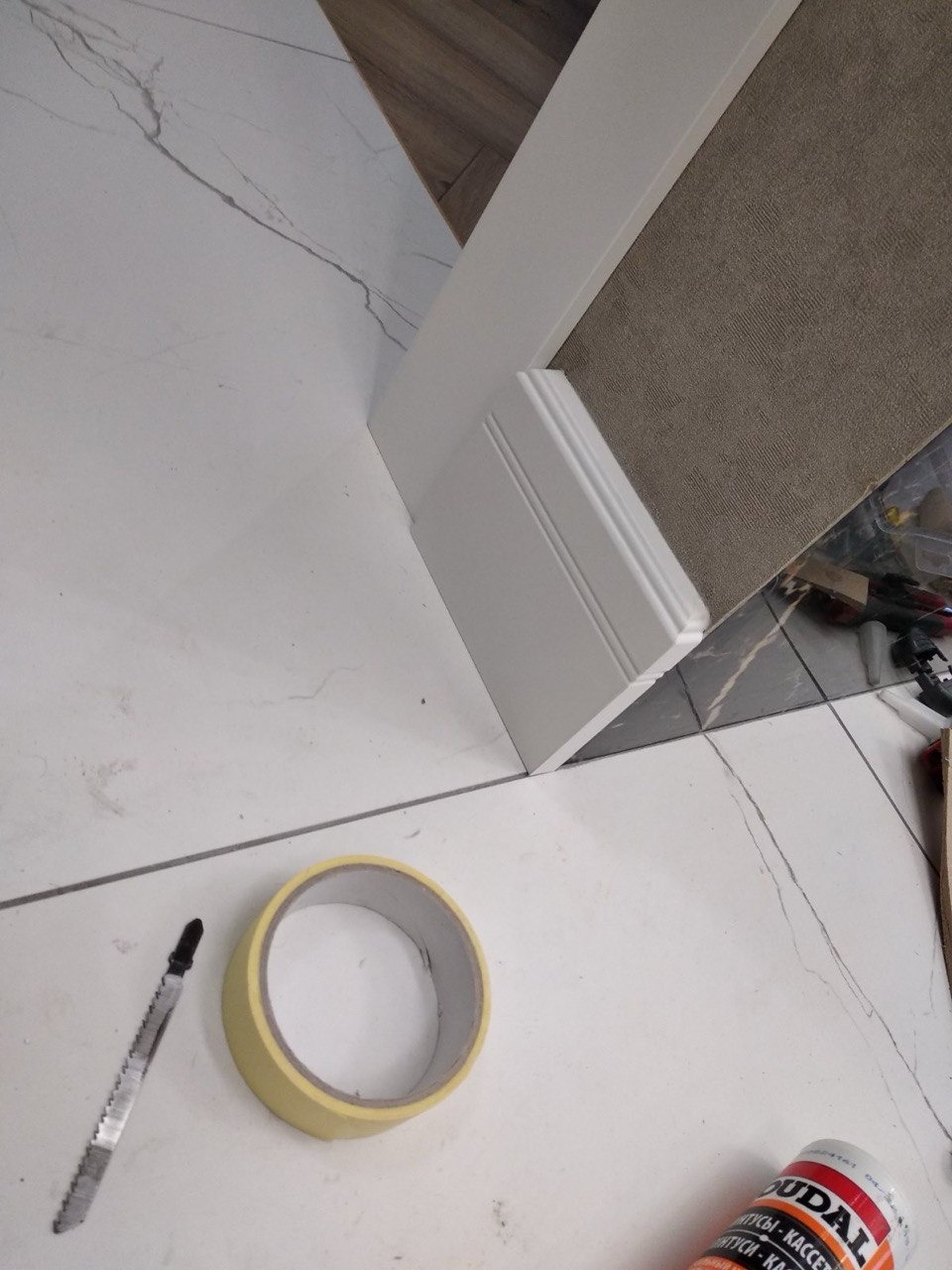 Połączenie podłogi z płytek imitujących marmur i paneli, z zamontowaną białą listwą przypodłogową, w tle narzędzia i materiały montażowe: taśma malarska, brzeszczot do wyrzynarki i tuba kleju.