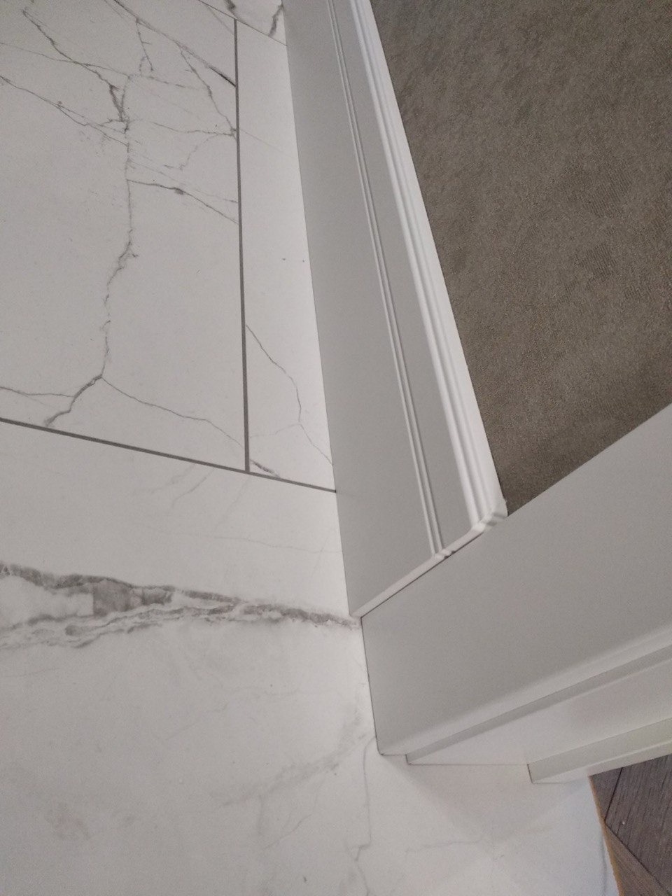 Połączenie podłogi wyłożonej białymi płytkami imitującymi marmur z szarą wykładziną dywanową, oddzielone białą listwą przypodłogową z frezami.