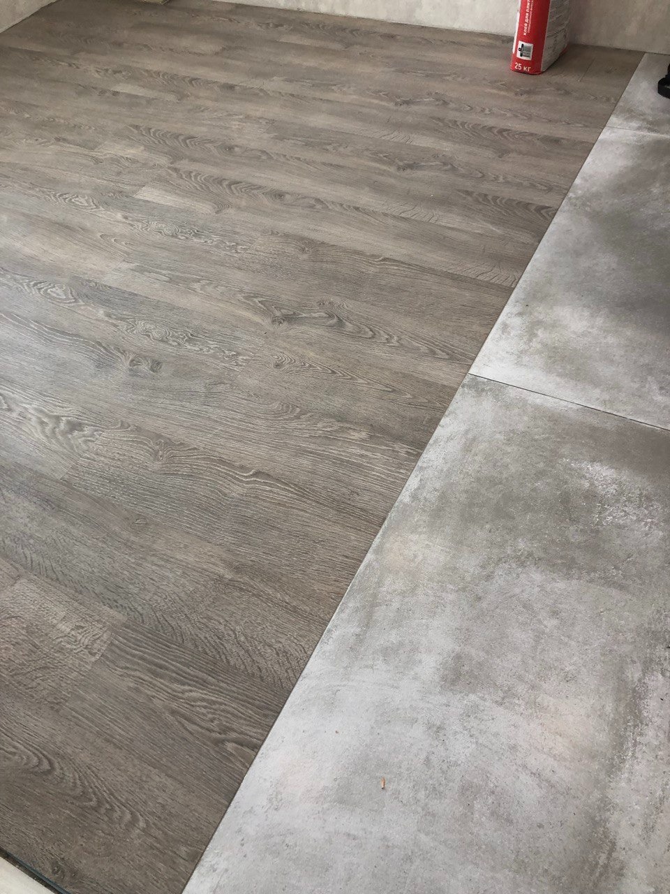 Połączenie szarych paneli podłogowych imitujących drewno z szarą posadzką wylewaną, widoczny worek z klejem w rogu pomieszczenia.