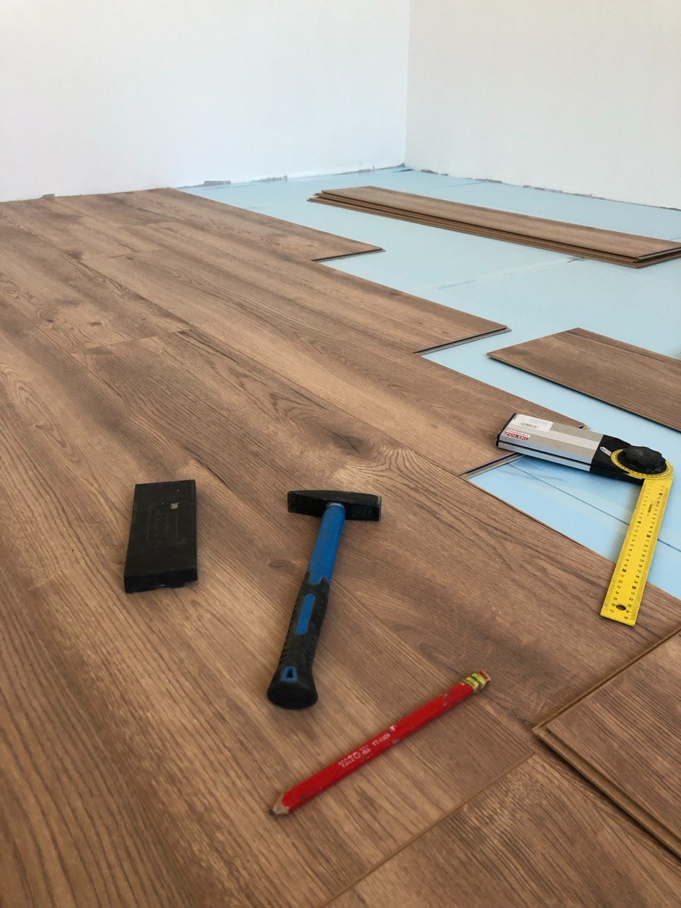 Fragment podłogi w trakcie montażu paneli w Warszawie, widoczne narzędzia: młotek z niebieskim trzonkiem, kątownik, ołówek stolarski i dobijak, pod spodem podkład w kolorze niebieskim.