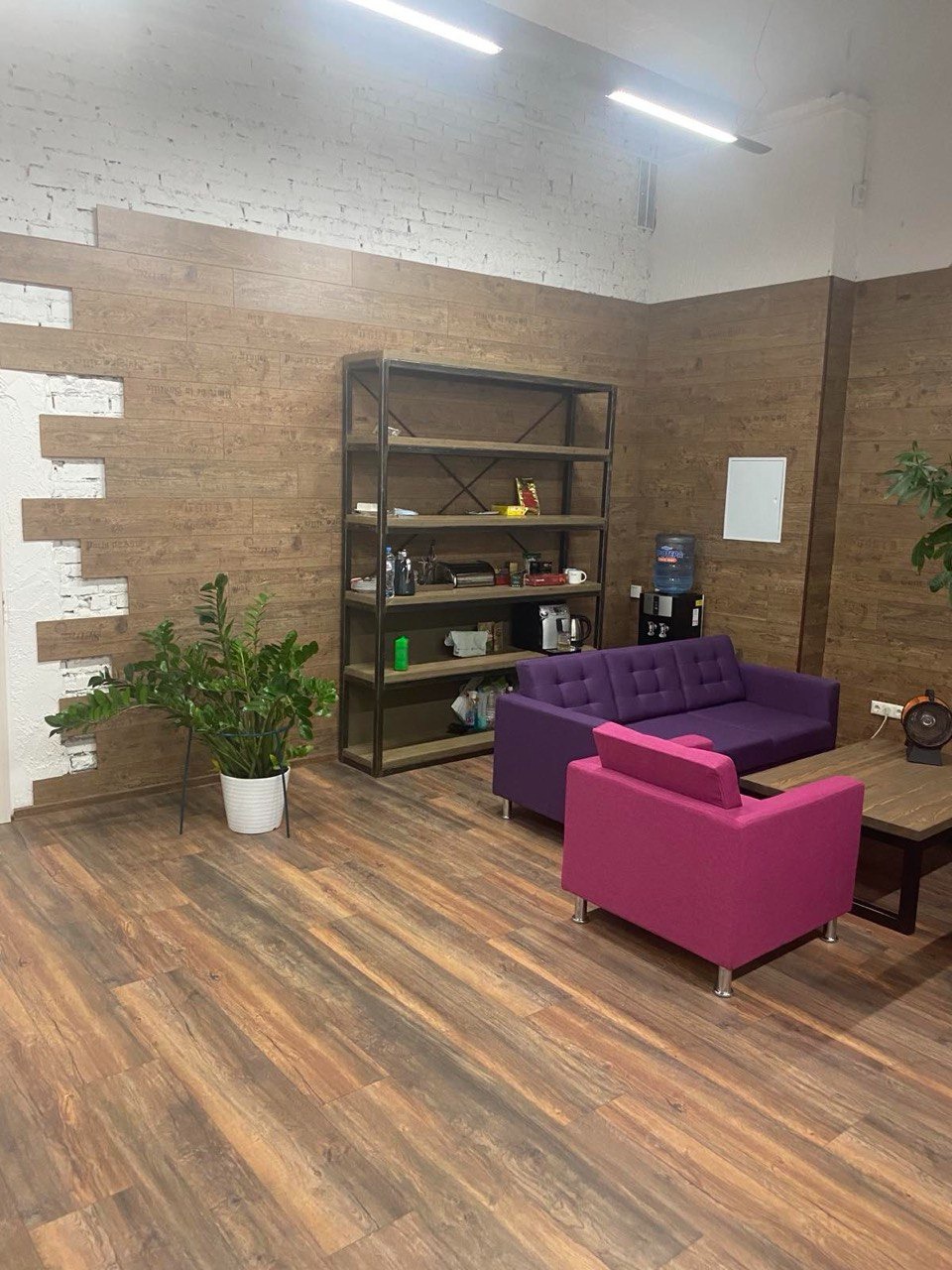 Nowoczesne wnętrze biurowe z drewnopodobną podłogą, fioletową sofą i różowym fotelem na tle ściany z elementami białej cegły i drewnianej okładziny.