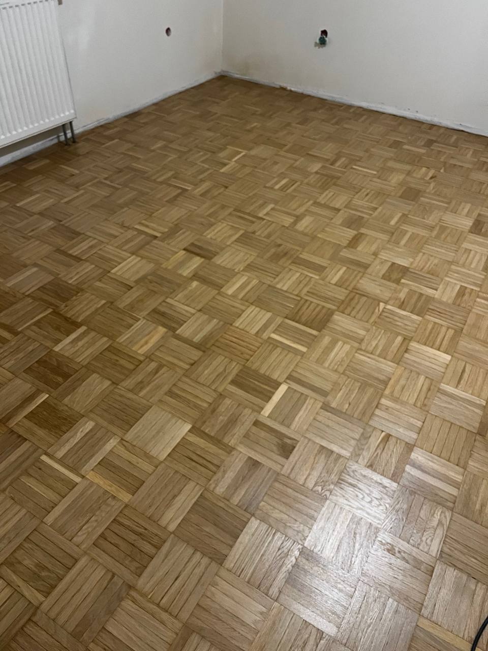 Odnowiona podłoga parkietowa w jodełkę, po cyklinowaniu i lakierowaniu. Widoczne naturalne usłojenie drewna i ciepła kolorystyka. W tle biała ściana z grzejnikiem.