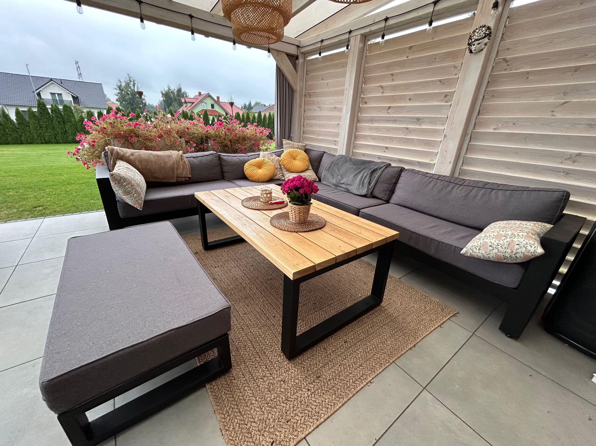Zadaszony taras z szarą sofą narożną, drewnianym stolikiem kawowym z czarnymi nogami, pufą i dywanem z juty, ozdobiony kwiatami i dekoracjami, oświetlony wiszącymi lampami.