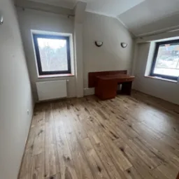 Pusty pokój na poddaszu z drewnianą podłogą, biurkiem i dwoma oknami z widokiem na zimowy krajobraz.