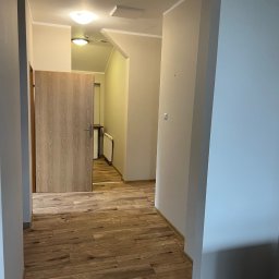 Haratyk - Korytarz z panelami podłogowymi w odcieniach brązu, kremowe ściany i drewniane drzwi z widocznym fragmentem grzejnika w przejściu.