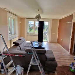 Haratyk - Salon w trakcie malowania, widoczne drabiny, brązowa sofa, okna z widokiem na zewnątrz, ściany w odcieniach beżu i brązu, podłoga z paneli imitujących drewno.