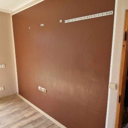 Haratyk - Czekoladowa ściana z widocznymi gniazdkami i listwą montażową, świeżo po malowaniu, z drewnopodobną podłogą i białym wykończeniem sufitu.