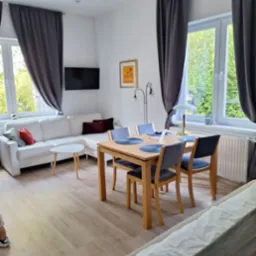 Jasne wnętrze pokoju dziennego z drewnianą podłogą, białym narożnikiem, stołem z szarymi krzesłami i szarymi zasłonami w oknach. Na ścianie wisi telewizor i obrazek.