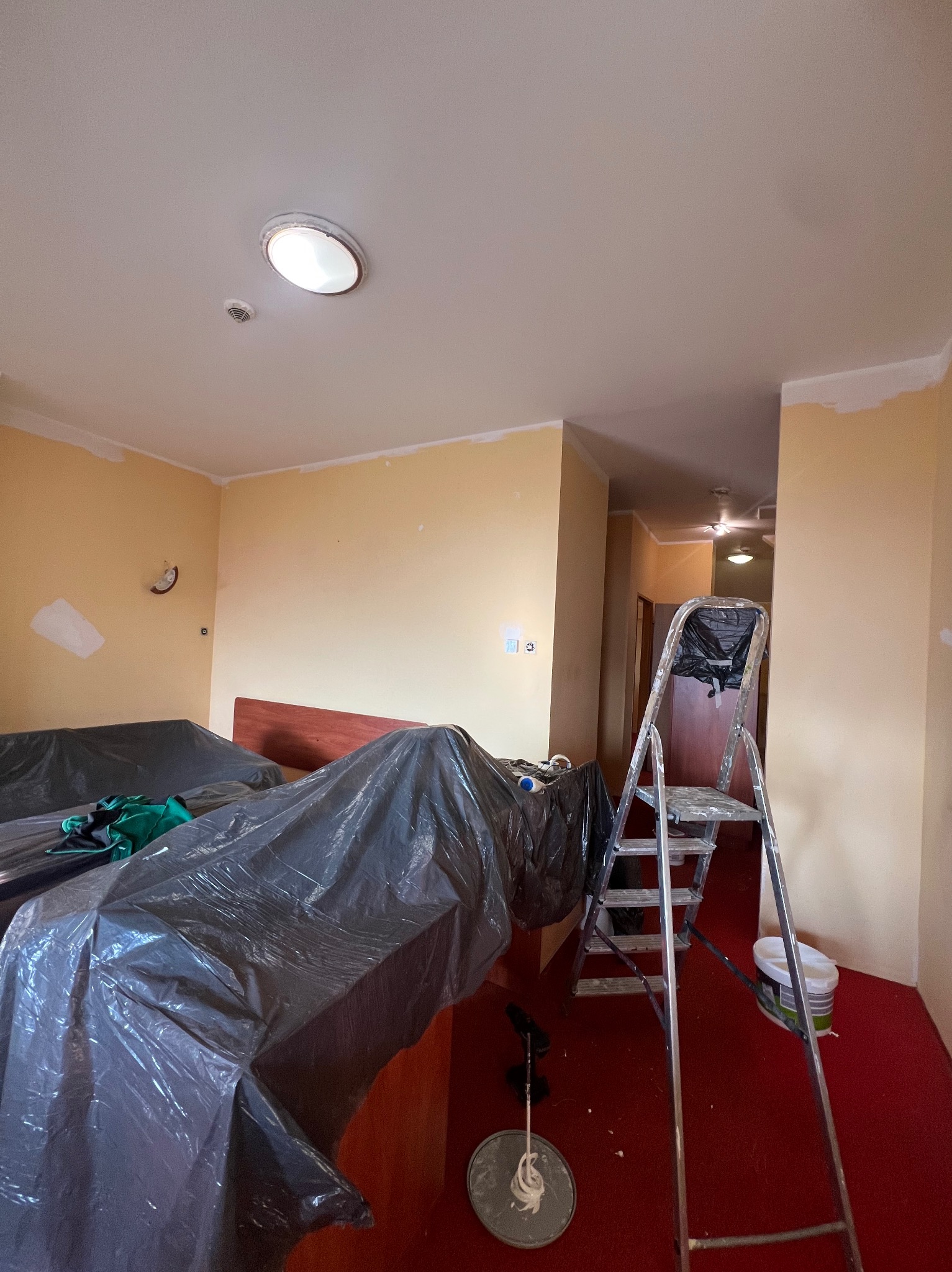 Pokój w trakcie malowania: meble zabezpieczone folią, drabina aluminiowa, puszka z farbą na czerwonej wykładzinie, ściany w odcieniach żółci z widocznymi fragmentami zagruntowanymi na biało.