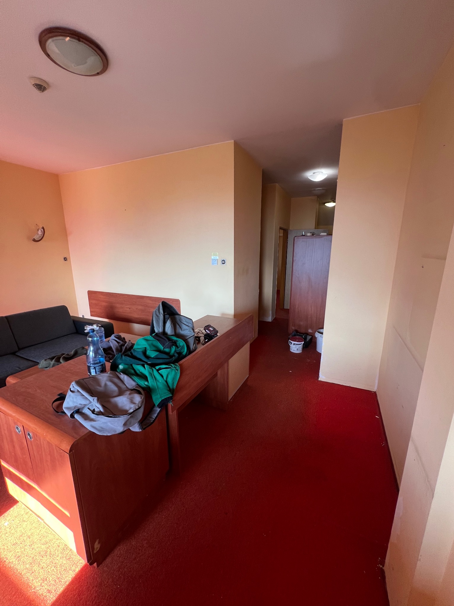 Pokój z czerwonym dywanem, beżowymi ścianami i meblami w trakcie remontu, widoczne farby i osobiste rzeczy na biurku.