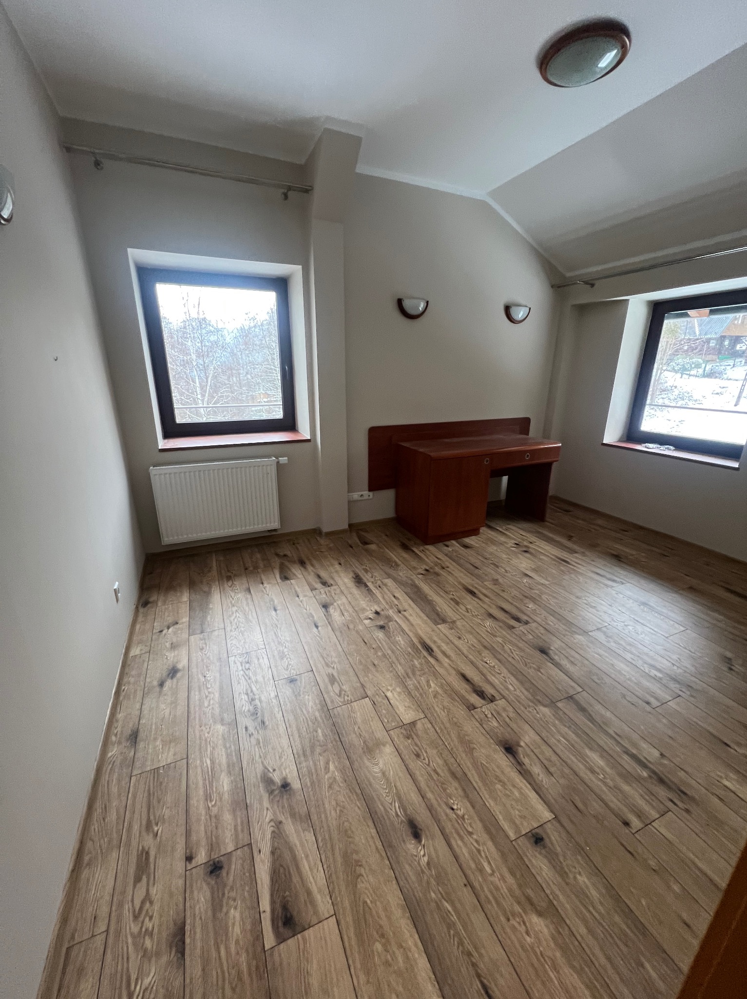 Pusty pokój na poddaszu z drewnianą podłogą, biurkiem i dwoma oknami z widokiem na zimowy krajobraz.