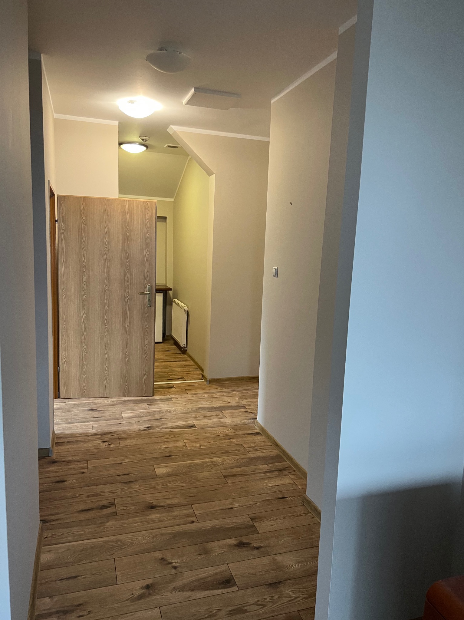 Korytarz z panelami podłogowymi w odcieniach brązu, kremowe ściany i drewniane drzwi z widocznym fragmentem grzejnika w przejściu.