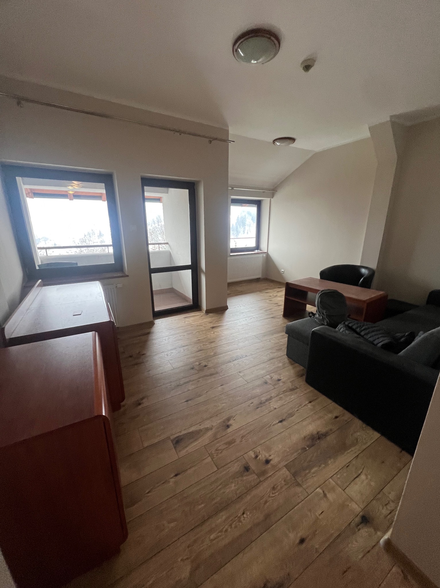 Jasne, przestronne wnętrze pokoju w Wiśle z drewnianą podłogą, widokiem z okna i wyjściem na balkon, częściowo umeblowane.