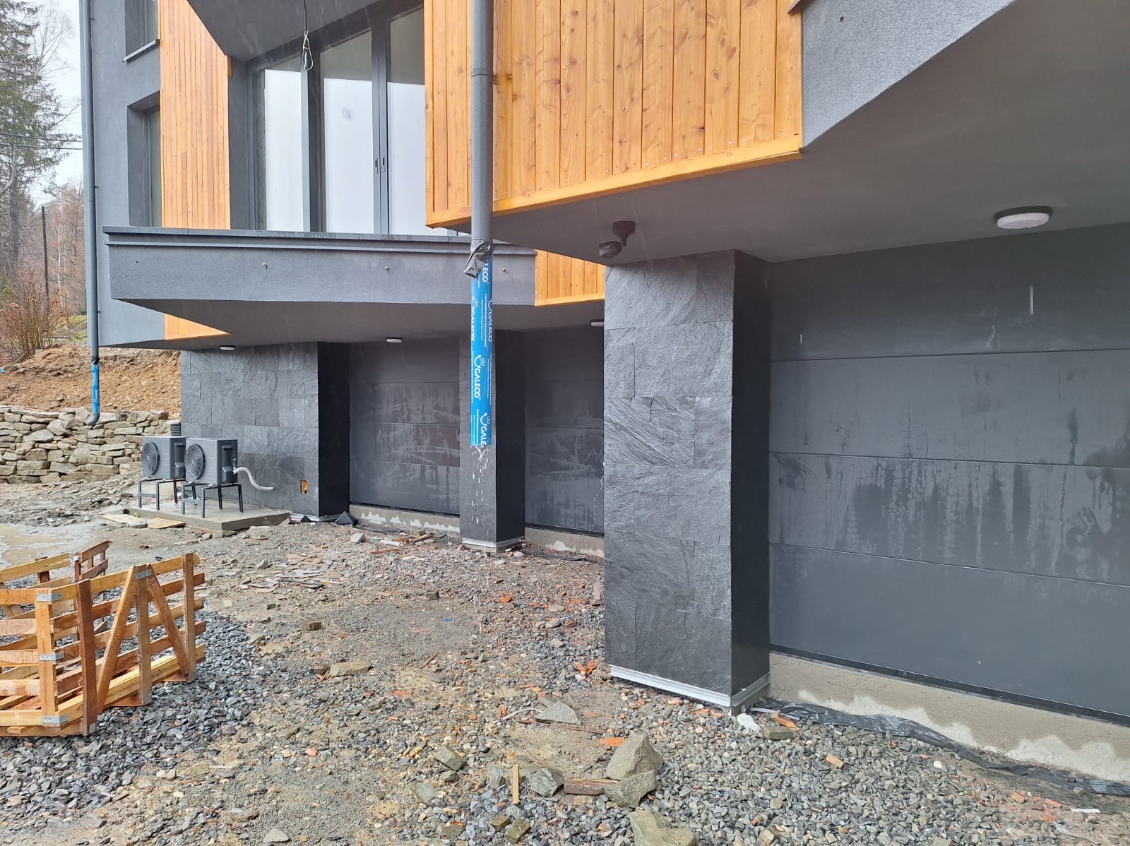 Elewacja budynku z szarego kamienia i drewnianymi elementami, widoczne dwa klimatyzatory na zewnątrz, teren wokół w trakcie prac wykończeniowych.